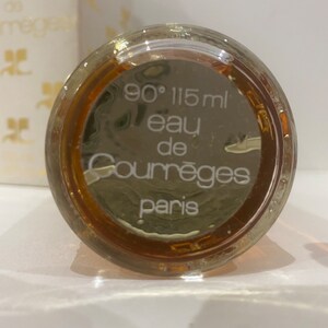 May include: The bottom of a clear glass bottle with a brown liquid inside. The bottle is labeled with white text that reads "90&deg; 115 ml eau de Courr&egrave;ges Paris".