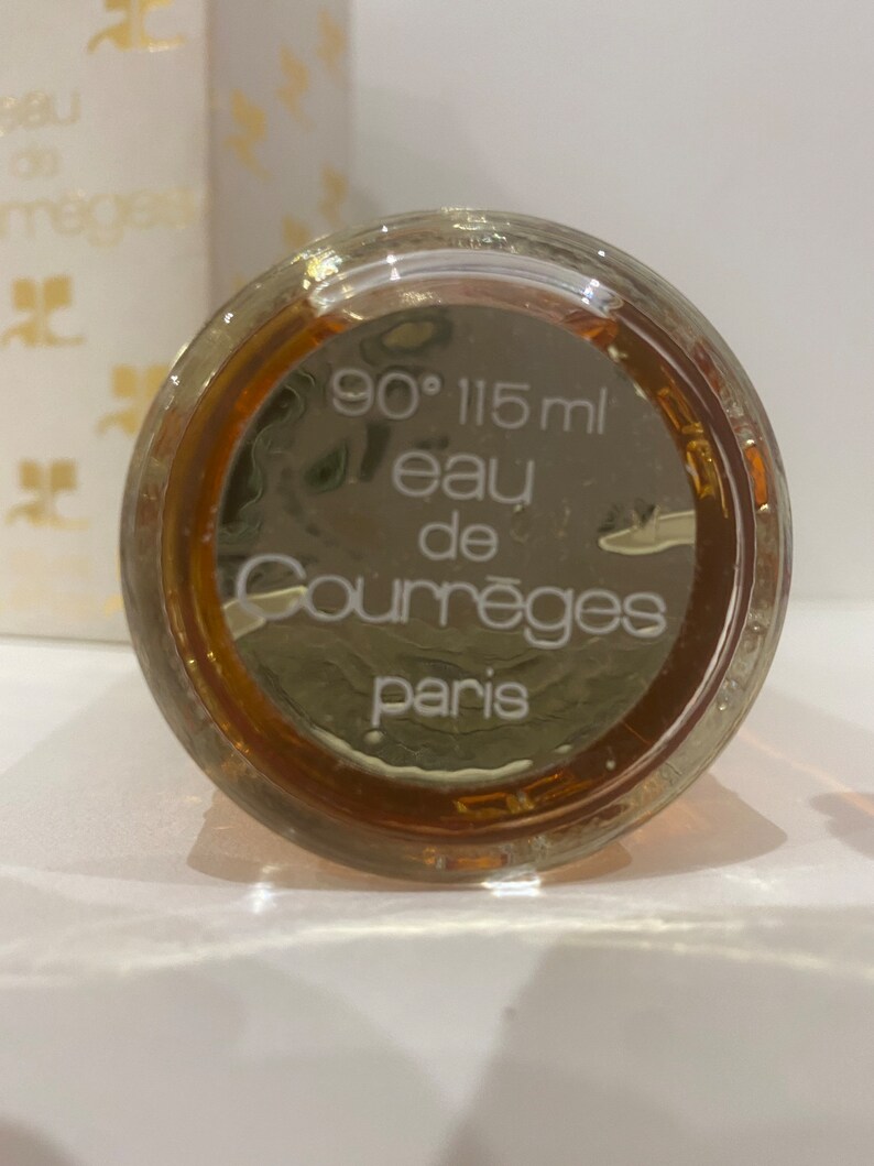 May include: The bottom of a clear glass bottle with a brown liquid inside. The bottle is labeled with white text that reads "90&deg; 115 ml eau de Courr&egrave;ges Paris".