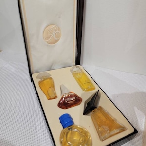 Puede incluir: Caja rectangular abierta con marco marrón oscuro e interior crema. Contiene seis frascos de perfume en miniatura de varias formas, llenos de líquido color ámbar. El interior está forrado con tela crema y tiene un emblema circular.