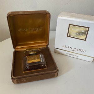 May include: A small, clear glass bottle of perfume with a gold cap and label. The bottle is in a brown velvet box with a gold label that reads "Jean Patou Paris". The box is next to a white box with a gold label that reads "Parfum "1000"" Jean Patou Paris 15 ml-e-1/2 fl. oz."
