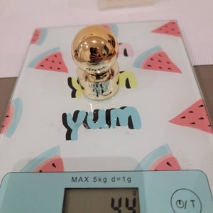 May include: A gold-colored bottle stopper on a digital scale with a watermelon print. The scale reads 44 grams.