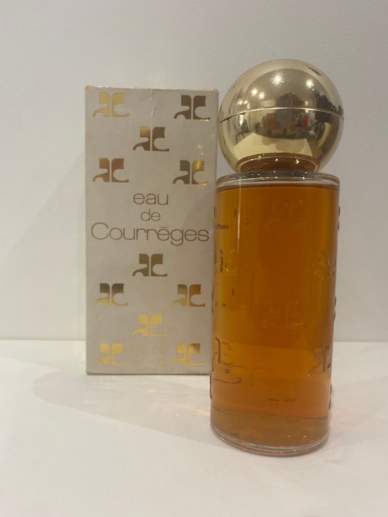May include: A clear glass bottle of Eau de Courr&egrave;ges perfume with a gold ball stopper. The bottle is filled with a light amber liquid. The bottle is sitting on a white surface. The box for the perfume is white with gold lettering and a repeating pattern of the letter "C".
