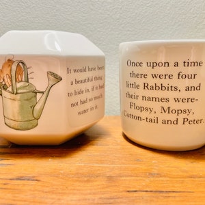 May include: Two white ceramic mugs with illustrations and text. The first mug is octagonal and features a rabbit holding a watering can. The text reads "It would have been a beautiful thing to hide in, if it had not had so much water in it." The second mug is round and features a rabbit with the text "Once upon a time there were four little Rabbits, and their names were- Flopsy, Mopsy, Cotton-tail and Peter."