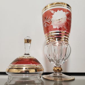 May include: A clear glass lidded jar and a tall stemmed glass goblet, both featuring deep red bands, intricate etched white floral patterns, and horizontal gold trim accents. The goblet has a fluted base and a gold-accented stem.