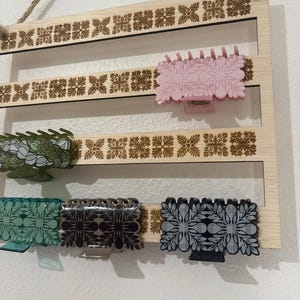May include: A light-colored wooden wall-mounted organizer with horizontal slats featuring laser-cut floral patterns. Five rectangular hair claw clips in various colors, including pink, green, teal, brown, and black, are attached to the slats.