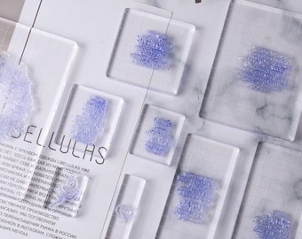 Könnte beinhalten: Ein Set mit acht transparenten Acrylstempeln mit einem Rastermuster. Die Stempel zeigen verschiedene Designs, darunter Herzen, Blumen und Wörter. Die Stempel sind perfekt, um Ihren Bastelprojekten eine persönliche Note zu verleihen.