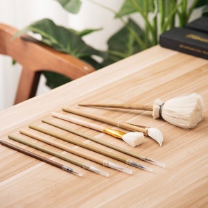 Puede incluir: Un conjunto de siete pinceles con mangos de madera y cerdas blancas. Los pinceles están dispuestos sobre una mesa de madera.