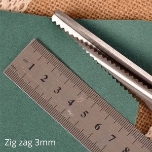 Puede incluir: Primer plano de unas tijeras en zigzag cortando un trozo de papel verde. Las tijeras est&aacute;n cortando un patr&oacute;n en zigzag de 3 mm. Se puede ver una regla en la imagen, que muestra la medida del patr&oacute;n en zigzag.