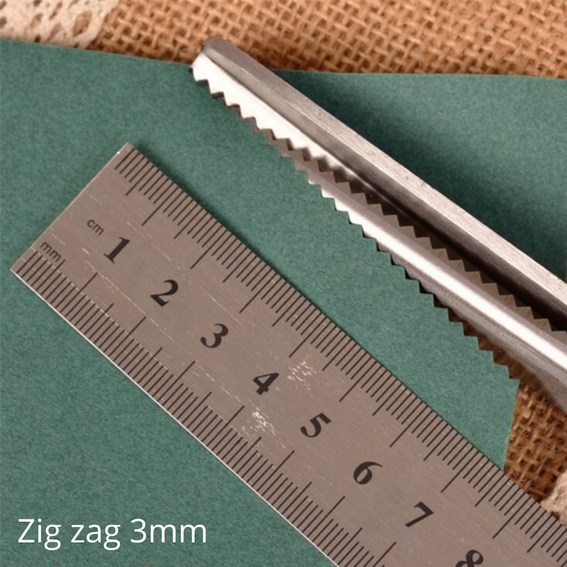 Puede incluir: Primer plano de unas tijeras en zigzag cortando un trozo de papel verde. Las tijeras est&aacute;n cortando un patr&oacute;n en zigzag de 3 mm. Se puede ver una regla en la imagen, que muestra la medida del patr&oacute;n en zigzag.