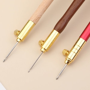 Op de afbeelding: Drie borduurnaalden met houten handvatten. De naalden hebben gouden metalen punten en een knop aan de bovenkant van het handvat. De handvatten zijn van naturel hout, bruin hout en rood.
