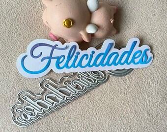 Puede incluir: Una plantilla de metal troquelada con la palabra "Felicidades" en un degradado azul y morado. La plantilla está sobre un fondo blanco con una pequeña figura de cerdo rosa.