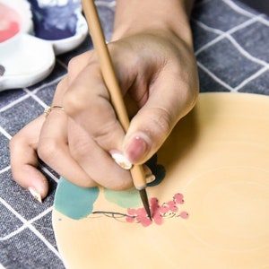 Puede incluir: Una mano sosteniendo un pincel de madera, pintando un dise&ntilde;o de flores rojas en un plato de cer&aacute;mica. El plato est&aacute; decorado con dise&ntilde;os de acuarela verde y azul.