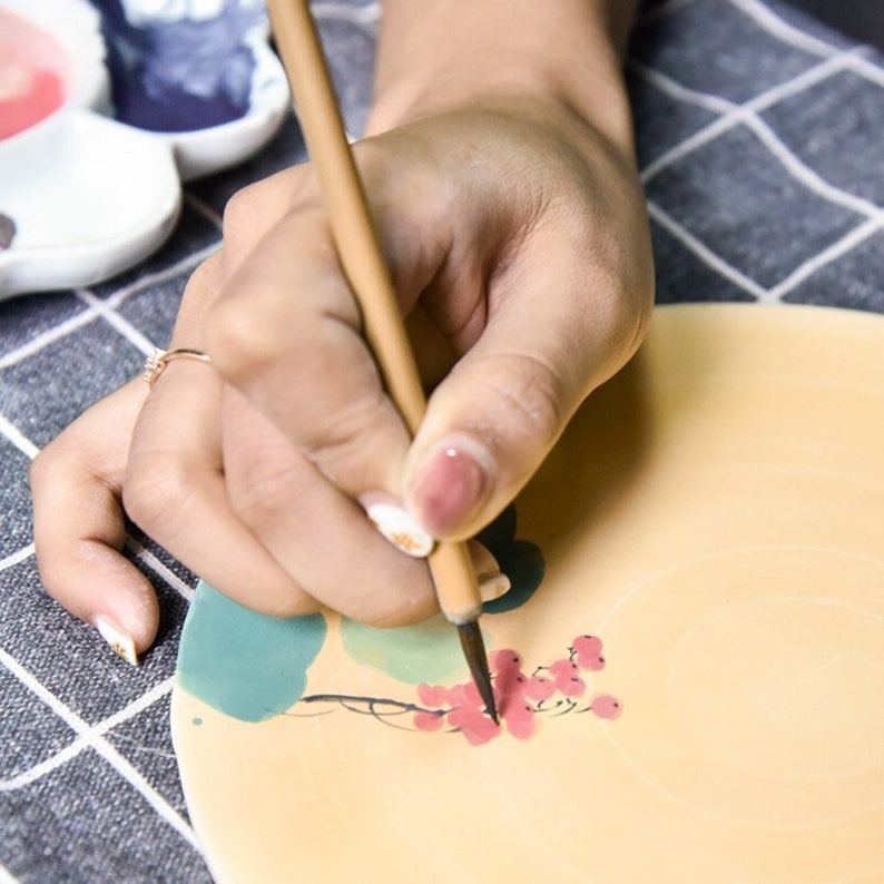 Puede incluir: Una mano sosteniendo un pincel de madera, pintando un dise&ntilde;o de flores rojas en un plato de cer&aacute;mica. El plato est&aacute; decorado con dise&ntilde;os de acuarela verde y azul.