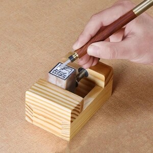 May include: A wooden stamp holder with a small, square, gray stamp with Chinese characters. A person is using a small, wooden-handled tool to carve the stamp.