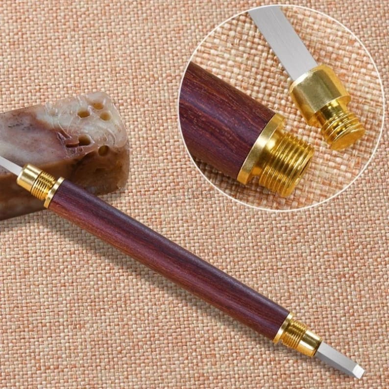 May include: A close-up of a wooden handled tool with a small, sharp blade. The tool has a gold-colored metal tip and a threaded end. The tool is lying on a brown fabric surface.