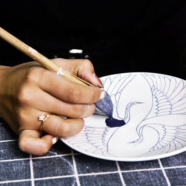 Puede incluir: Una persona est&aacute; pintando un plato de cer&aacute;mica blanco con un pincel azul. El plato tiene un dise&ntilde;o de dos grullas con las alas extendidas.