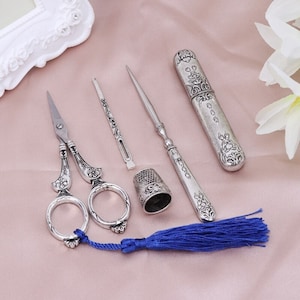 Op de afbeelding: Een zilveren naaiset met een schaar, een vingerhoed, een naaldinvoerder, een naald en een naaldkussen. De set wordt geleverd met een blauwe kwast.