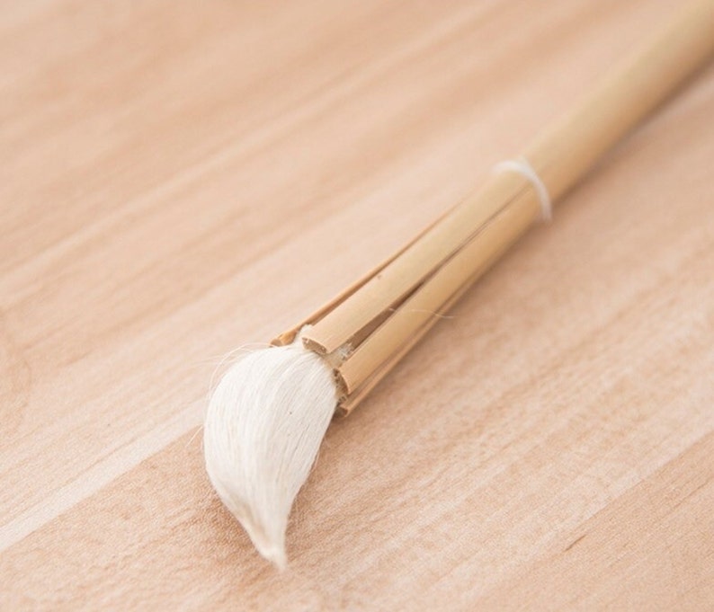 Puede incluir: Un pincel de caligraf&iacute;a blanco con un mango de bamb&uacute; natural. El pincel est&aacute; sobre una superficie de madera marr&oacute;n claro.