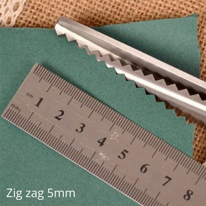 Puede incluir: Primer plano de unas tijeras de zig zag plateadas cortando un trozo de tela verde. Se ve una regla con marcas en cent&iacute;metros y mil&iacute;metros, que muestra que la puntada de zig zag tiene 5 mm de ancho.
