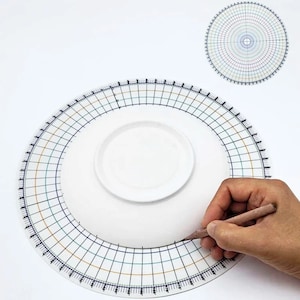 May include: A white ceramic plate with a black and gold grid pattern around the edge. The plate is being used as a drawing surface with a pencil.