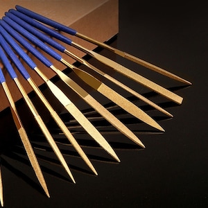 Puede incluir: Un juego de 12 limas de metal de color dorado con mangos azules. Las limas están dispuestas en forma de abanico, con los mangos apuntando hacia la parte superior de la imagen. Las limas se utilizan para dar forma y alisar el metal.