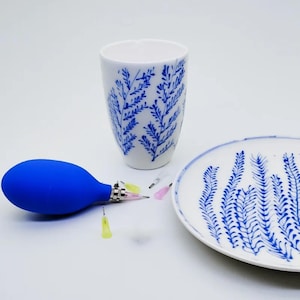 May include: A white ceramic cup and plate with blue leaf patterns. A blue bulb syringe with needles is next to the cup and plate. The cup is cylindrical, and the plate is round.