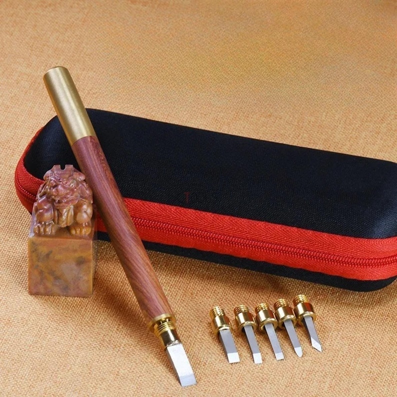 May include: A set of six small carving tools with wooden handles and metal blades, in a black zippered case. The tools are arranged in a row, with the handles pointing to the right. The case is closed, and the zipper is pulled up. The tools are used for carving wood, leather, and other materials.