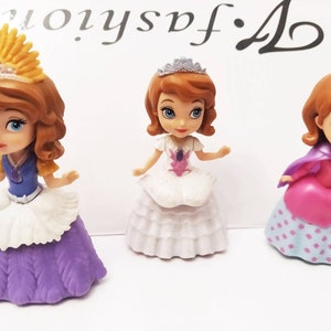 Puede incluir: Tres pequeñas muñecas de plástico de una princesa con cabello castaño que llevan vestidos diferentes. La princesa de la izquierda lleva un vestido morado con un delantal blanco y una corona amarilla. La princesa del medio lleva un vestido blanco con un diseño de flores moradas. La princesa de la derecha lleva un vestido rosa con una falda de lunares azules y blancos y una capa morada.