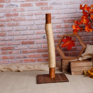 May include: A wooden cat scratching post with a natural rope covering. The post is on a wooden base and is in front of a brick wall.
