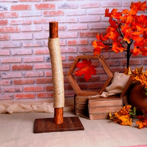 May include: A wooden cat scratching post with a natural rope covering. The post is on a brown base and is in front of a brick wall. There are fall-themed decorations around the post, including a wooden hexagon frame with a red maple leaf inside, a brown box, and a brown fabric.