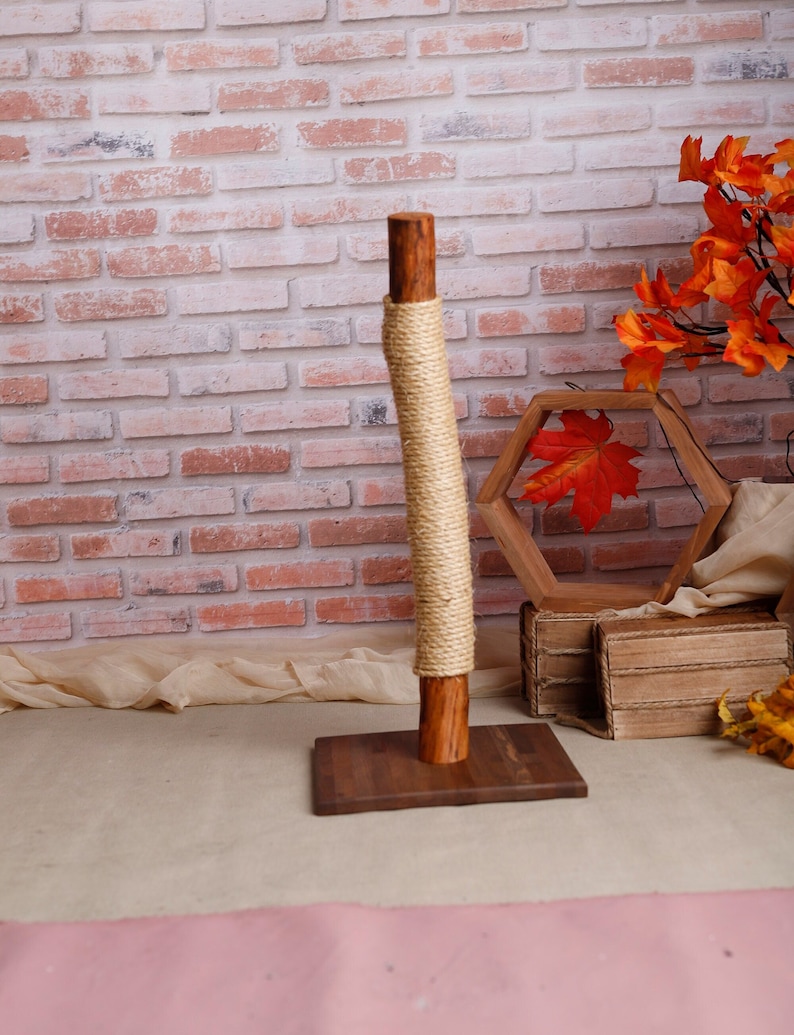 May include: A wooden cat scratching post with a natural rope covering. The post is on a wooden base and is in front of a brick wall.