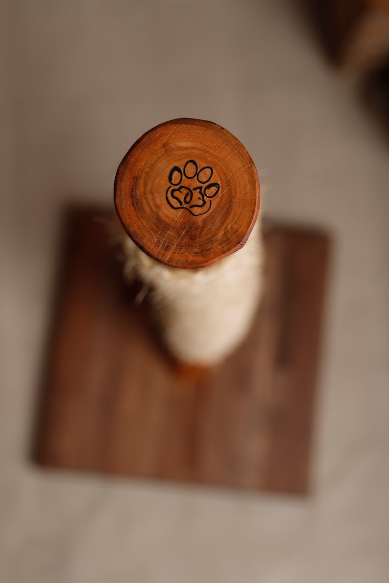 May include: A wooden cat scratching post with a paw print design on the top. The post is wrapped in sisal rope and is mounted on a wooden base.