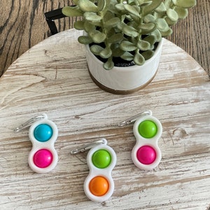 Op de afbeelding: Drie witte, dubbelzijdige dimple fidget-speeltjes met een karabijnhaak. Elk speeltje heeft twee felgekleurde siliconen bellen: blauw en roze, groen en oranje en groen en roze. Een kleine plant in een pot staat op de achtergrond.