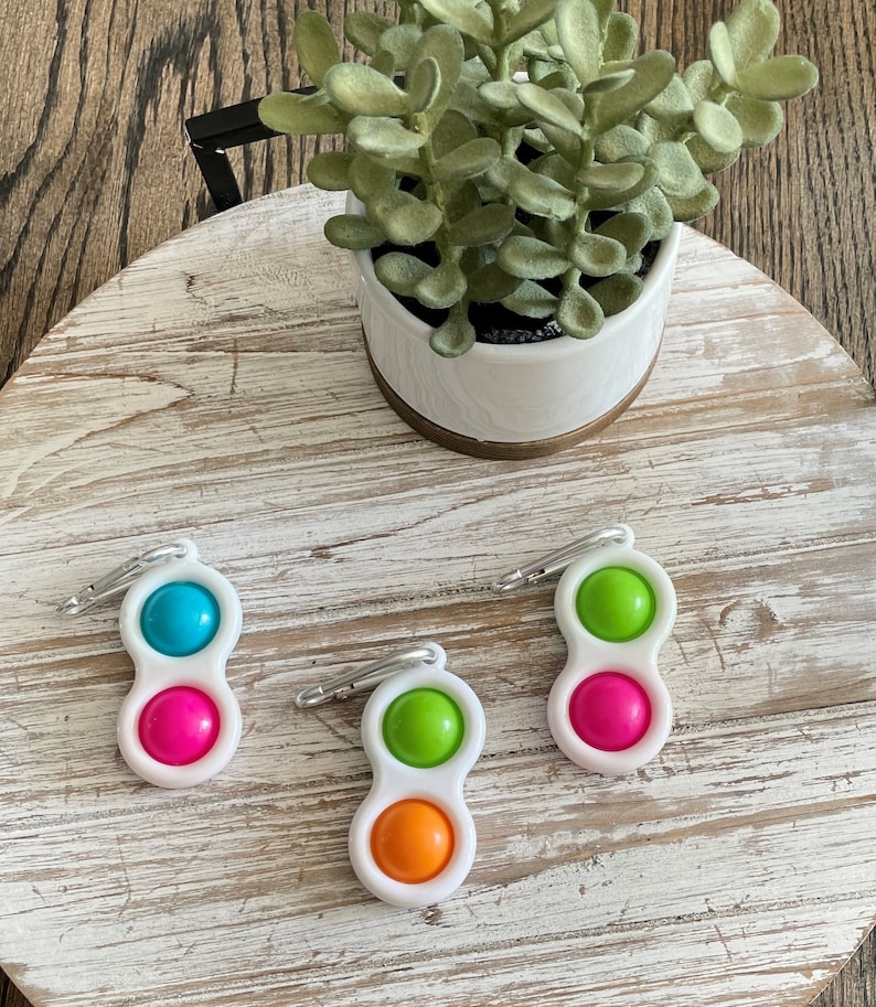 Op de afbeelding: Drie witte, dubbelzijdige dimple fidget-speeltjes met een karabijnhaak. Elk speeltje heeft twee felgekleurde siliconen bellen: blauw en roze, groen en oranje en groen en roze. Een kleine plant in een pot staat op de achtergrond.