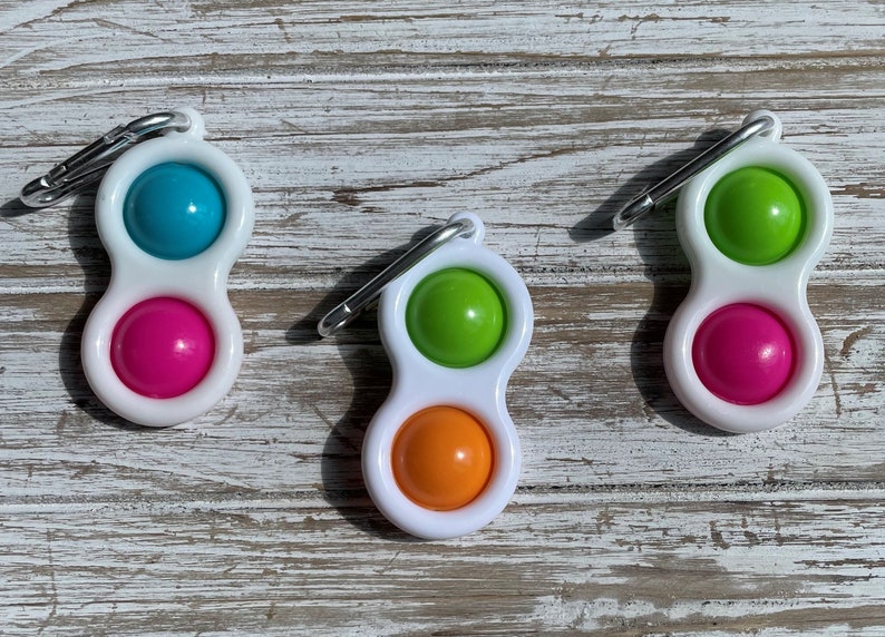 Op de afbeelding: Drie witte fidget-speeltjes met een karabijnhaak. Elk speeltje heeft twee gekleurde siliconen bellen: blauw en roze, groen en oranje, en groen en roze. De speeltjes liggen op een verweerd, wit houten oppervlak.