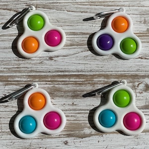 May include: Four white dimple fidget toys with colorful spheres in green, orange, purple, blue, and pink. Each toy has a silver carabiner clip attached. The toys are arranged on a weathered, white wooden surface.