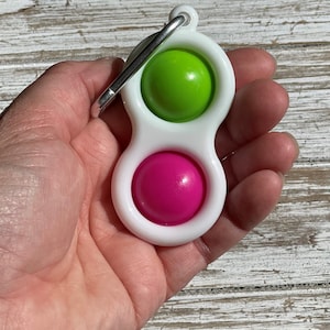 Op de afbeelding: Een wit plastic fidget-speeltje met een groene en roze bol. Het speelgoed heeft een zilveren metalen clip. Het speelgoed wordt in een hand gehouden.