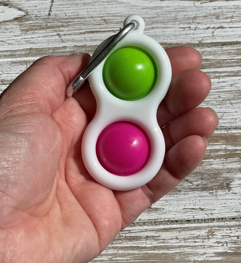 Op de afbeelding: Een wit plastic fidget-speeltje met een groene en roze bol. Het speelgoed heeft een zilveren metalen clip. Het speelgoed wordt in een hand gehouden.
