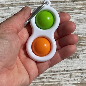 Op de afbeelding: Een wit plastic fidget-speeltje met een groen en oranje bubbelontwerp. Het speelgoed heeft een zilveren metalen clip aan de bovenkant. Het speelgoed wordt in een hand gehouden.