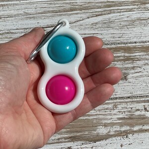 Op de afbeelding: Een wit plastic fidget-speeltje met een turquoise en roze bal aan elk uiteinde. Het speelgoed heeft een zilveren metalen clip aan de bovenkant bevestigd. Het speelgoed wordt in de hand van een persoon gehouden.