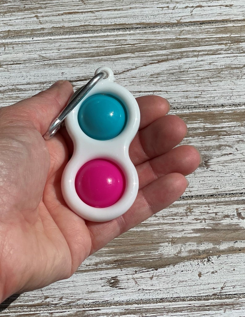 Op de afbeelding: Een wit plastic fidget-speeltje met een turquoise en roze bal aan elk uiteinde. Het speelgoed heeft een zilveren metalen clip aan de bovenkant bevestigd. Het speelgoed wordt in de hand van een persoon gehouden.
