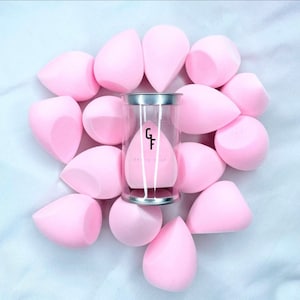 May include: A set of 13 pink makeup sponges in a clear container with a silver lid. The sponges are teardrop shaped and have a smooth texture. The container has the text "Gracefully" printed on it.