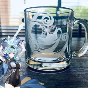 May include: A clear glass mug with a handle, featuring an etched design of swirling waves and a snowflake. The mug is on a dark surface, with a blurred background of a window and blinds.