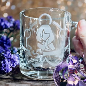 May include: Clear glass mug with a handle, etched with a white design featuring a stylized figure. The mug is on a wooden surface, with purple flowers in the background. The mug is held by a person.