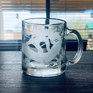 May include: A clear glass mug with a white etched design of a dragon. The mug has a handle and is sitting on a wooden surface.