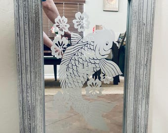 May include: Rectangular mirror with a distressed white wooden frame. The mirror features an etched design of a koi fish and floral accents. The frame has a weathered appearance, adding a rustic touch to the decor.