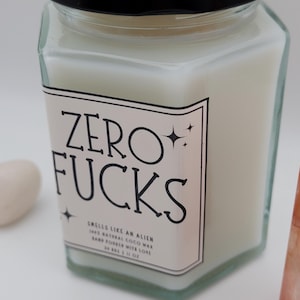 May include: A square glass jar candle with a black lid. The label reads "ZERO FUCKS" in black text. The candle is filled with a white wax and has a label that says "Smells like an Alien".