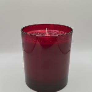 May include: A cylindrical, red candle in a glass container. The candle has a white wick and a glittery surface. The glass is a deep red colour, with a slightly darker base. The candle is unscented.
