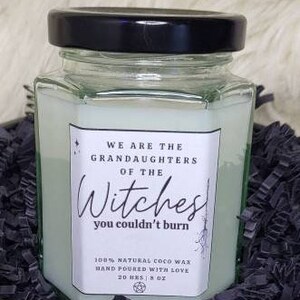 May include: A hexagonal glass jar candle with a black lid. The candle has a white label with the text "WE ARE THE GRANDDAUGHTERS OF THE Witches you couldn't burn". The candle is filled with a light green wax and is 227 grams.
