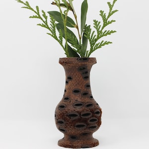 May include: A brown wooden vase with a unique design featuring a pattern of small, round holes. The vase is filled with green leafy branches.